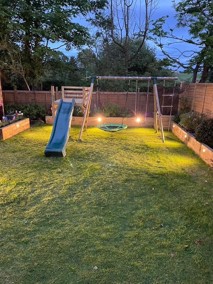 Family garden with a lit lawn, borders and play area after dark