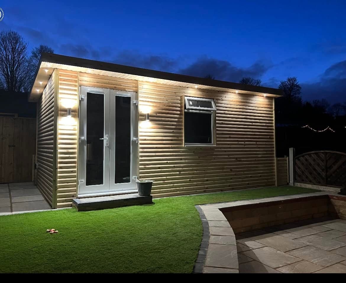 Cedar-clad garden room lit at dusk, with exterior wall lights and warm interior lighting through the doors